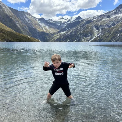 A young boy stands in the shallow, clear water of a mountain lake, posing with his hands raised and a playful expression. In the background, large, snow-dusted mountains rise under a partly cloudy blue sky.