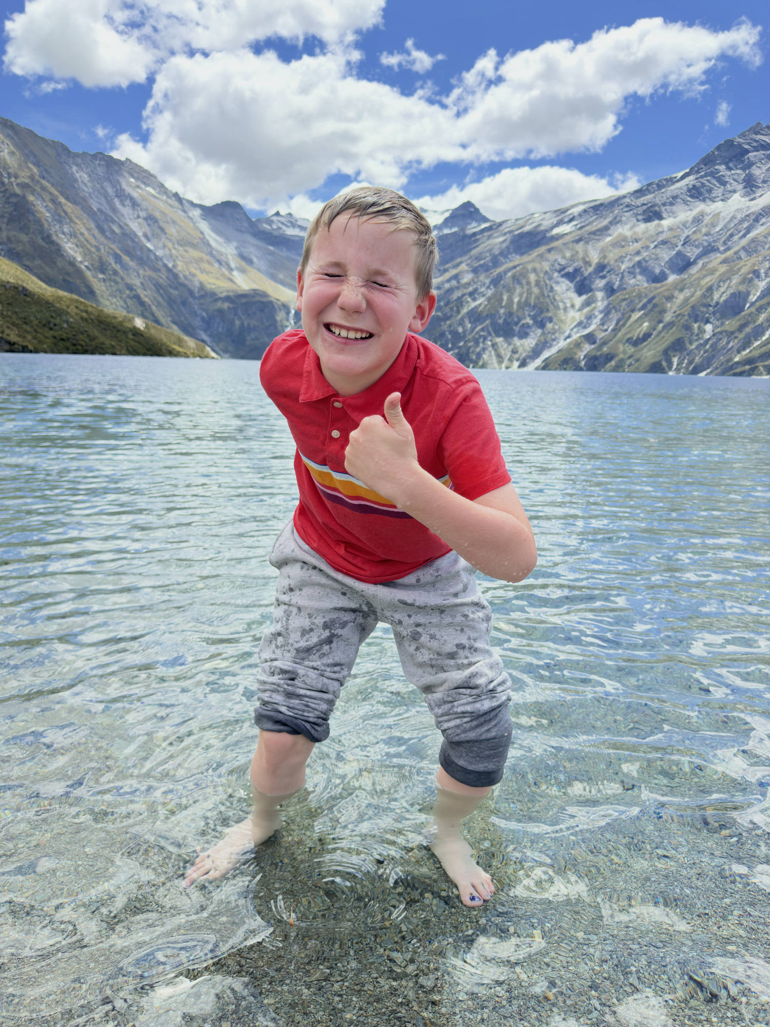 A young boy in a red shirt and grey pants stands in the shallow water of a clear mountain lake, giving a thumbs-up and squinting with a big smile. The background features a scenic landscape of large, rugged mountains under a partly cloudy blue sky.