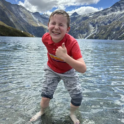 A young boy in a red shirt and grey pants stands in the shallow water of a clear mountain lake, giving a thumbs-up and squinting with a big smile. The background features a scenic landscape of large, rugged mountains under a partly cloudy blue sky.