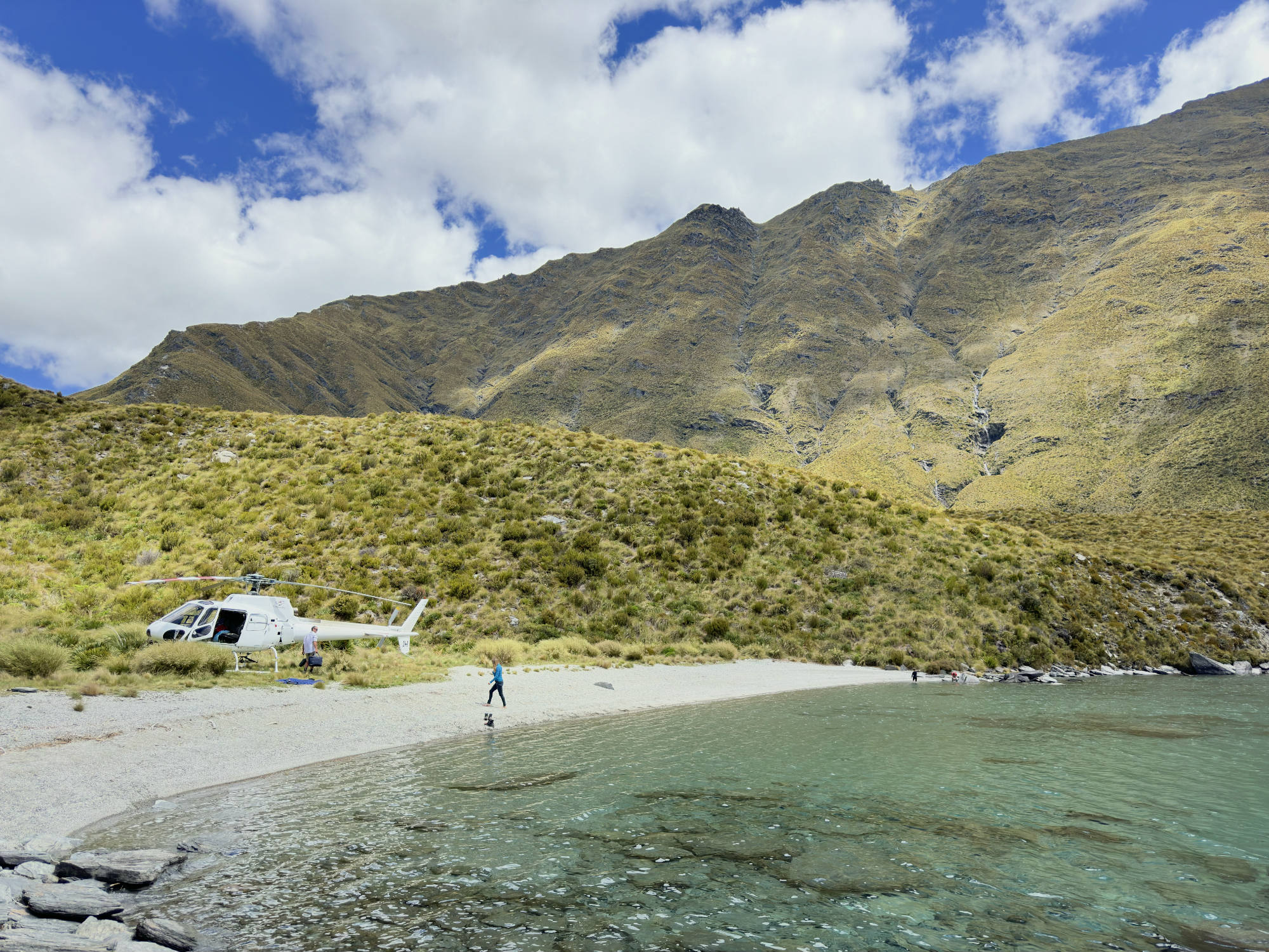 A white helicopter is parked on a gravel beach next to clear, shallow water, with a few people walking nearby. The scene is set in a mountainous landscape under a blue sky with white clouds.