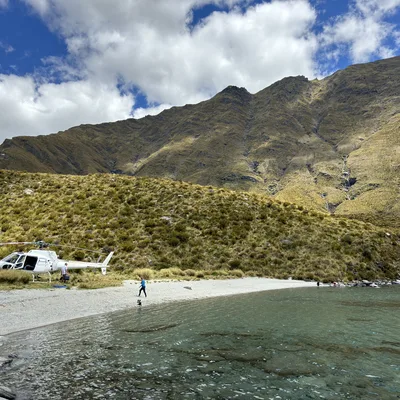 A white helicopter is parked on a gravel beach next to clear, shallow water, with a few people walking nearby. The scene is set in a mountainous landscape under a blue sky with white clouds.