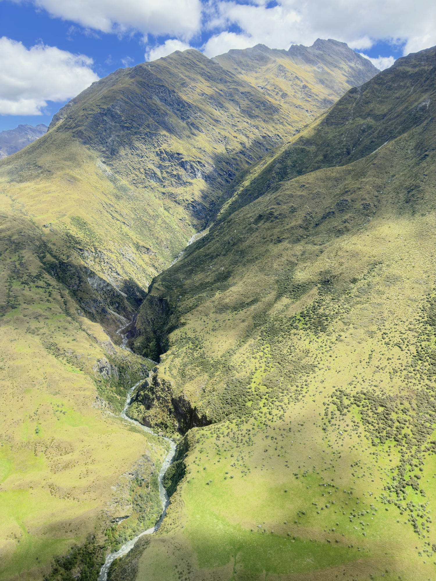 An aerial view captures a winding river flowing through a deep, grassy valley between steep mountain slopes under a partly cloudy sky. Shadows from the clouds are cast across the green and yellow terrain of the mountain landscape.