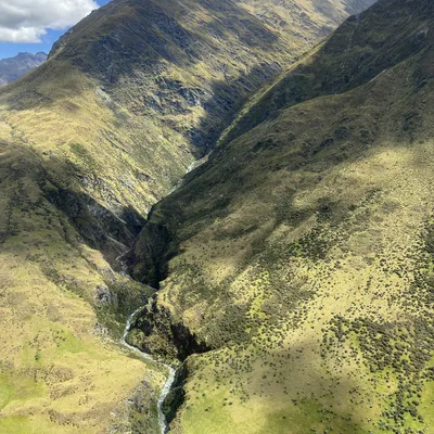 An aerial view captures a winding river flowing through a deep, grassy valley between steep mountain slopes under a partly cloudy sky. Shadows from the clouds are cast across the green and yellow terrain of the mountain landscape.