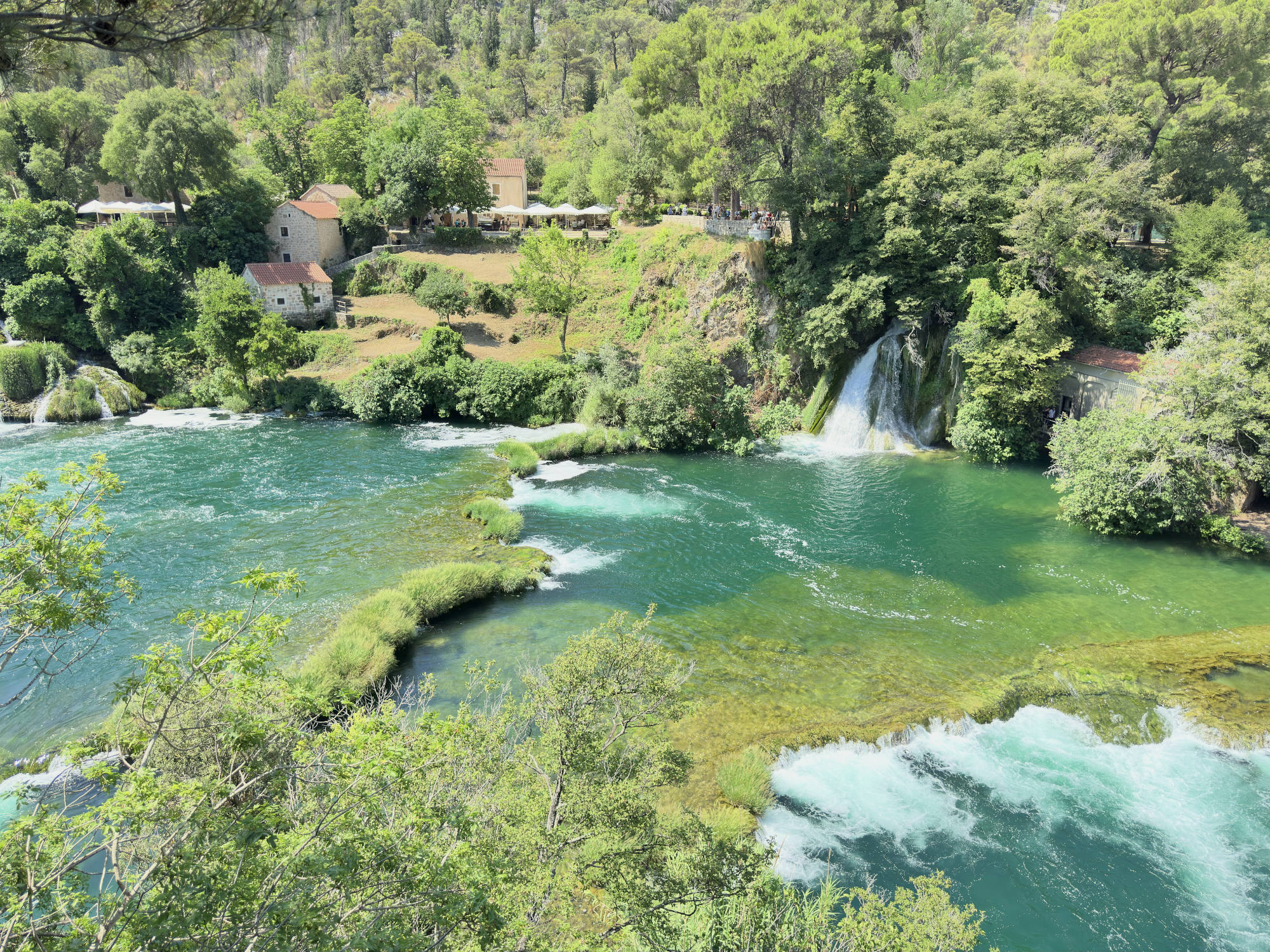 An elevated, wide shot captures a scenic river landscape featuring turquoise water flowing over travertine barriers and a small waterfall. Lush green trees and vegetation line the banks, with stone buildings and outdoor dining areas visible on the hillside in the background.