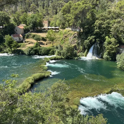 An elevated, wide shot captures a scenic river landscape featuring turquoise water flowing over travertine barriers and a small waterfall. Lush green trees and vegetation line the banks, with stone buildings and outdoor dining areas visible on the hillside in the background.