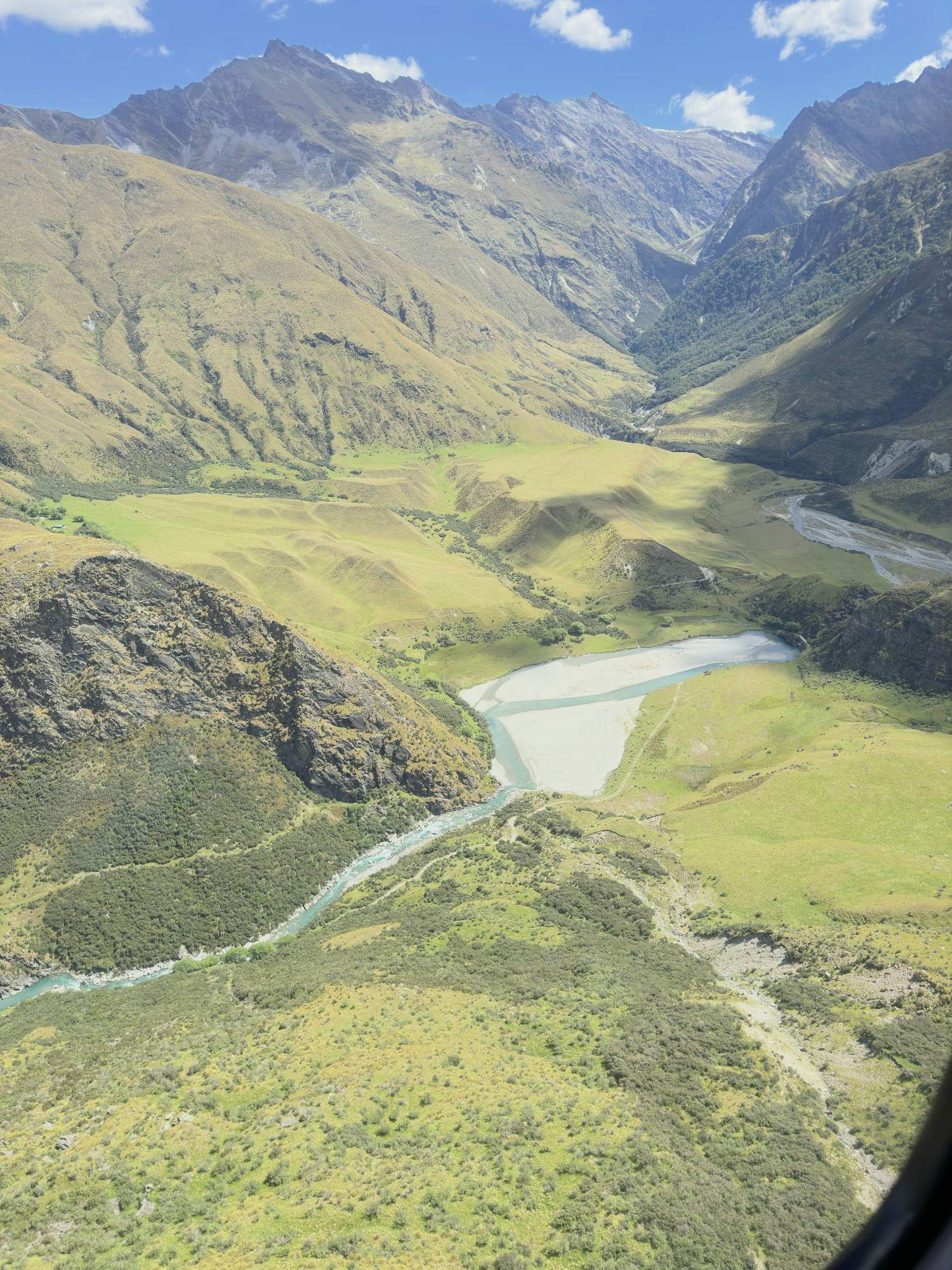 An aerial view captures a winding, light-blue river flowing through a lush green valley surrounded by steep, rugged mountains under a partly cloudy sky. The landscape features rolling grassy hills, rocky slopes, and a deep mountain pass in the background.