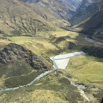 An aerial view captures a winding, light-blue river flowing through a lush green valley surrounded by steep, rugged mountains under a partly cloudy sky. The landscape features rolling grassy hills, rocky slopes, and a deep mountain pass in the background.