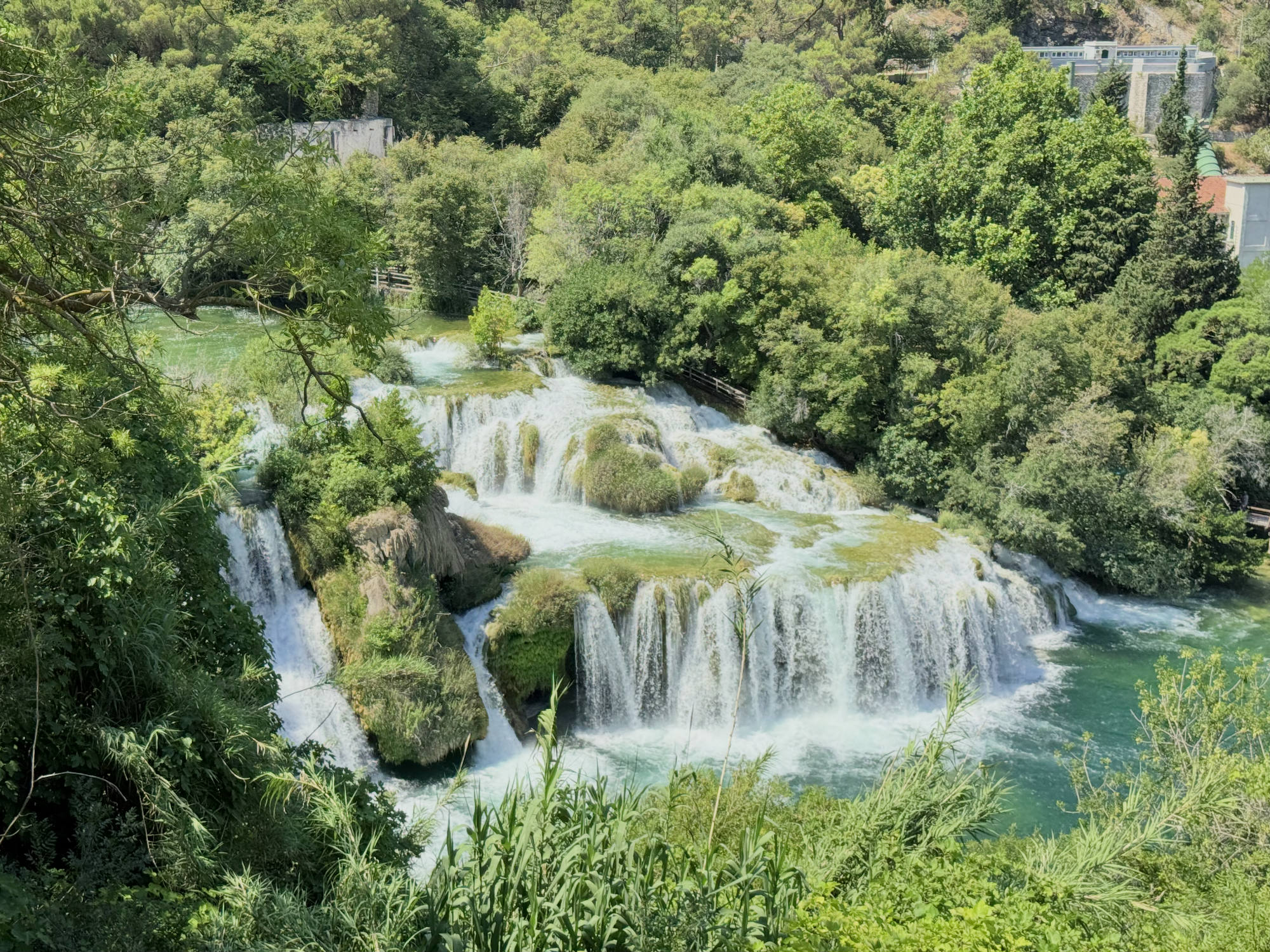 A high-angle shot captures a multi-tiered waterfall cascading over mossy rocks into a turquoise pool, surrounded by dense green forest. In the background, buildings are partially visible among the trees on the hillside.