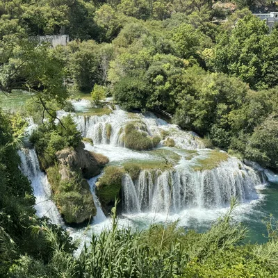 A high-angle shot captures a multi-tiered waterfall cascading over mossy rocks into a turquoise pool, surrounded by dense green forest. In the background, buildings are partially visible among the trees on the hillside.