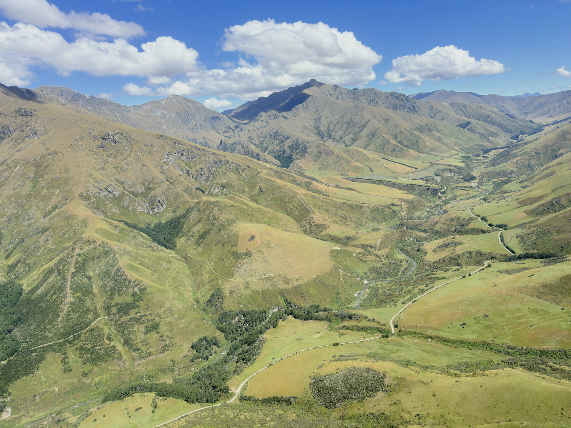 This high-angle aerial photograph captures a vast, rolling mountain range with green and brown slopes under a blue sky with fluffy white clouds. A winding dirt road and a small river snake through the valley below, highlighting the rugged, natural landscape.