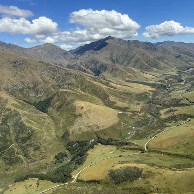 This high-angle aerial photograph captures a vast, rolling mountain range with green and brown slopes under a blue sky with fluffy white clouds. A winding dirt road and a small river snake through the valley below, highlighting the rugged, natural landscape.