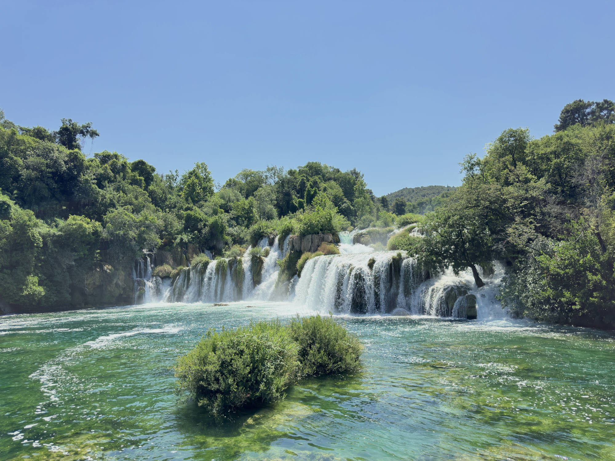 A wide waterfall cascades over rocky, tree-lined cliffs into a clear, turquoise pool of water under a bright blue sky. Green shrubs emerge from the water in the foreground, surrounded by lush forest vegetation on either side of the falls.