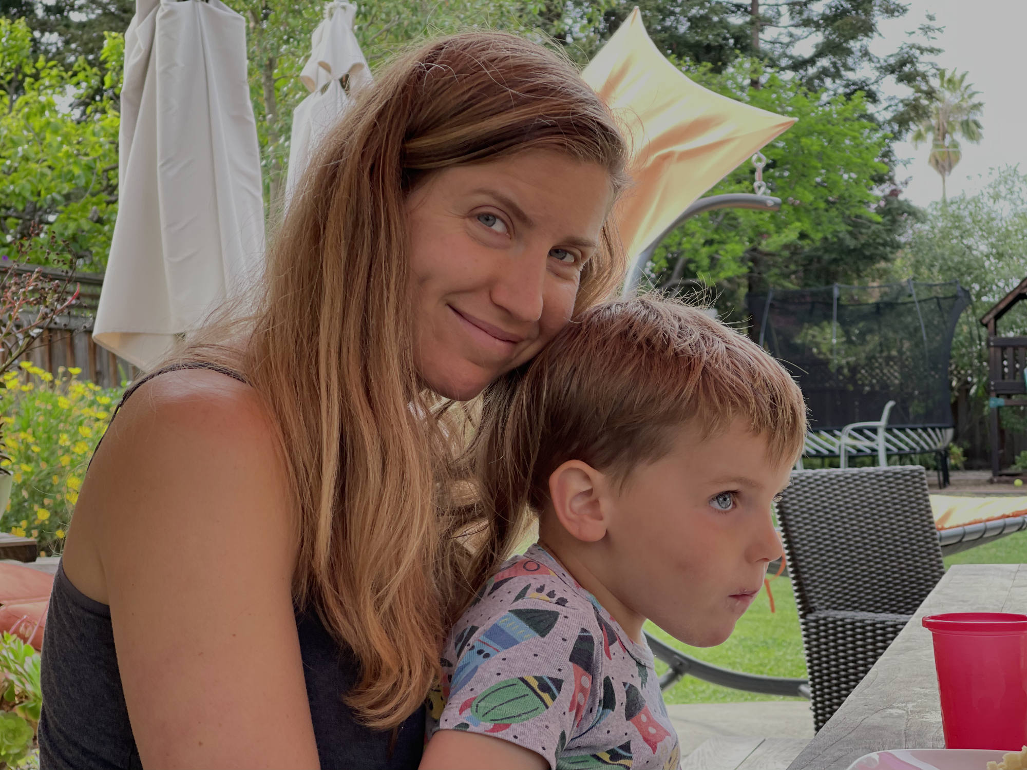 A smiling woman with long brown hair poses with her head resting against a young boy sitting in her lap at an outdoor patio table. The backyard setting features patio furniture, umbrellas, and green foliage in the background.