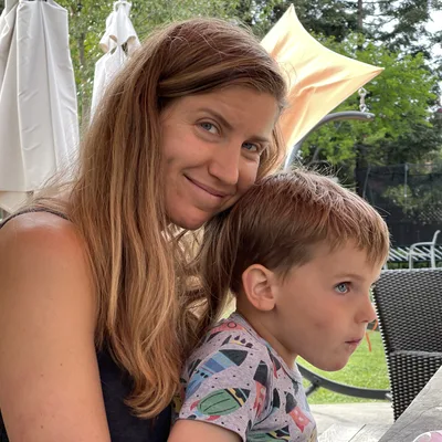 A smiling woman with long brown hair poses with her head resting against a young boy sitting in her lap at an outdoor patio table. The backyard setting features patio furniture, umbrellas, and green foliage in the background.