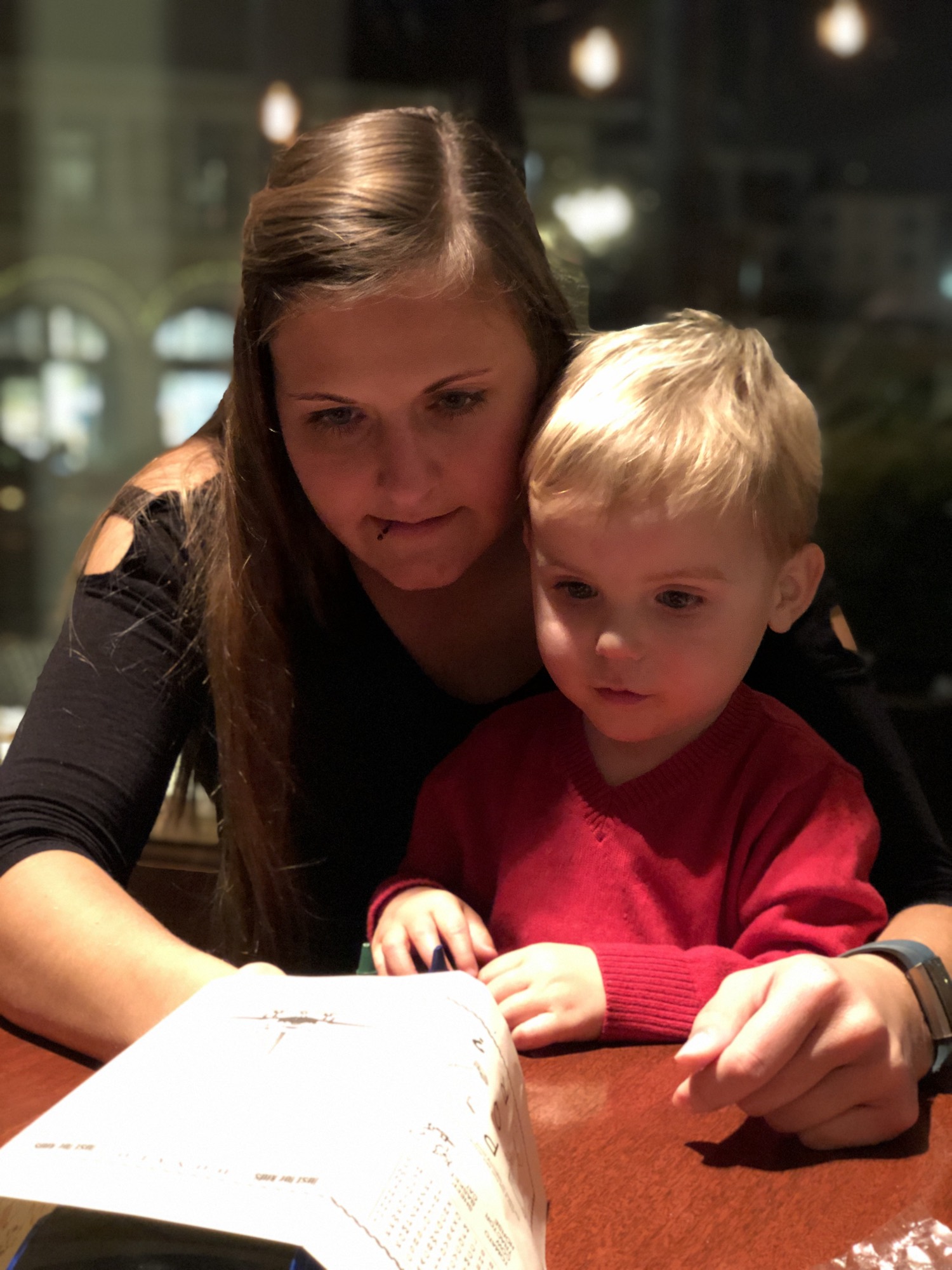 A woman in a black shirt and a young boy in a red sweater sit together at a wooden table, looking down at a paper activity menu. They are indoors, likely in a restaurant, with soft, warm lighting and a dark window in the background.