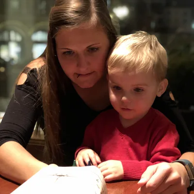 A woman in a black shirt and a young boy in a red sweater sit together at a wooden table, looking down at a paper activity menu. They are indoors, likely in a restaurant, with soft, warm lighting and a dark window in the background.