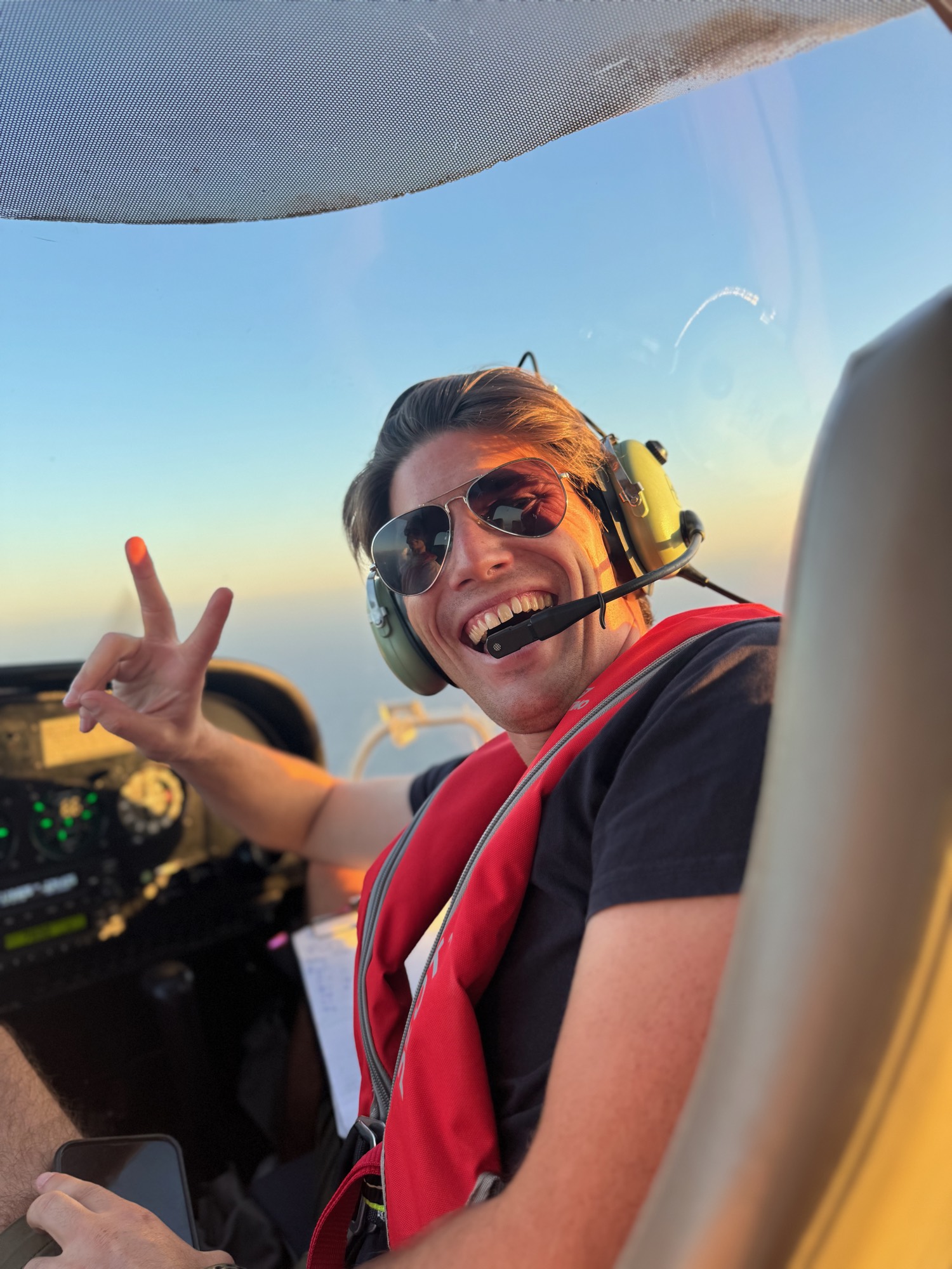 A smiling man wearing sunglasses, an aviation headset, and a red life vest flashes a peace sign from the cockpit of a small airplane. The photo is taken from inside the cabin during flight, with a clear blue sky visible through the windshield.