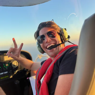 A smiling man wearing sunglasses, an aviation headset, and a red life vest flashes a peace sign from the cockpit of a small airplane. The photo is taken from inside the cabin during flight, with a clear blue sky visible through the windshield.