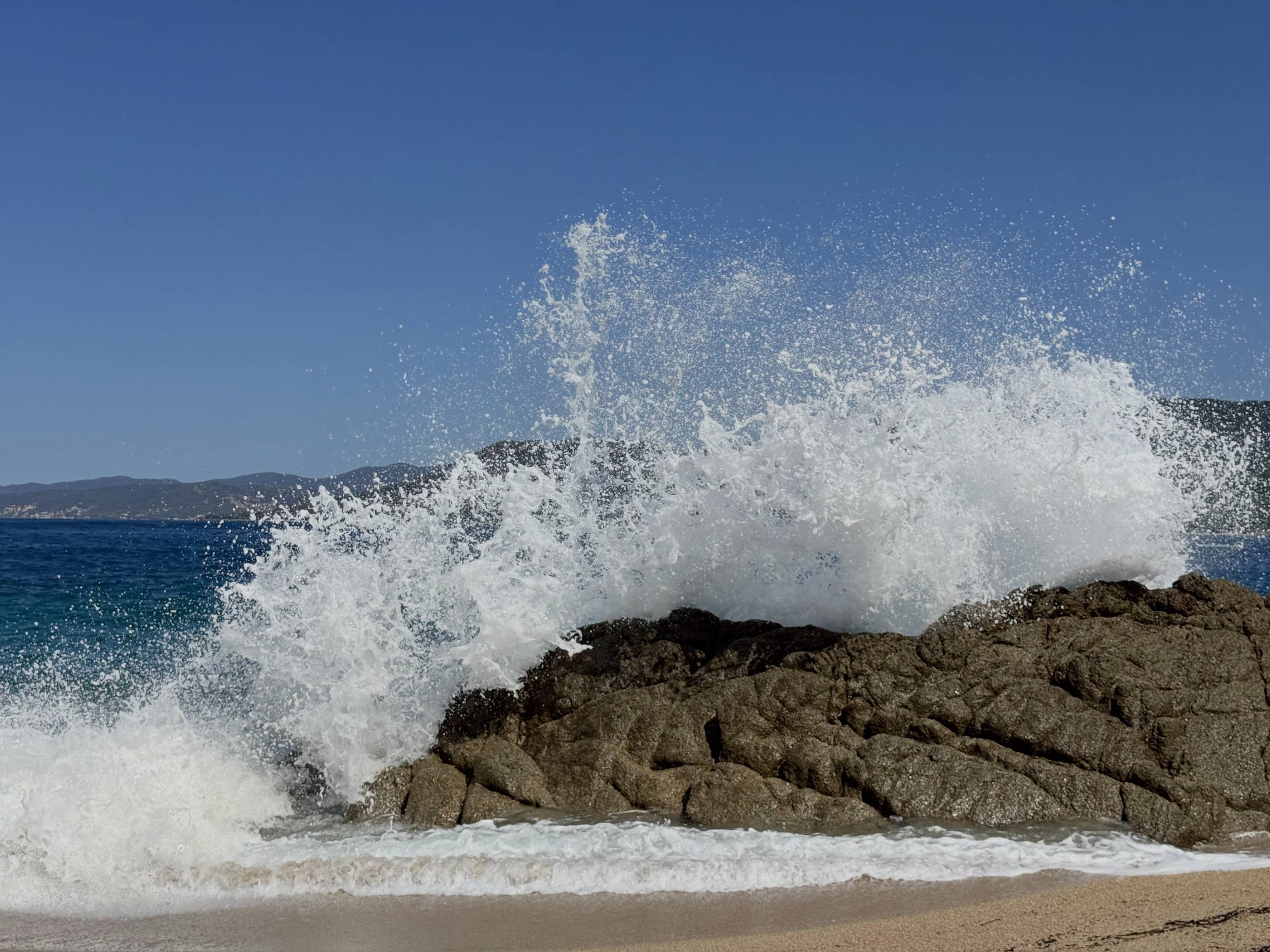 A large ocean wave crashes over a rocky outcrop on a sandy beach, creating a dramatic spray of white foam and water. The scene is set against a clear blue sky with distant hills visible across the water.