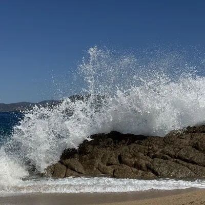 A large ocean wave crashes over a rocky outcrop on a sandy beach, creating a dramatic spray of white foam and water. The scene is set against a clear blue sky with distant hills visible across the water.