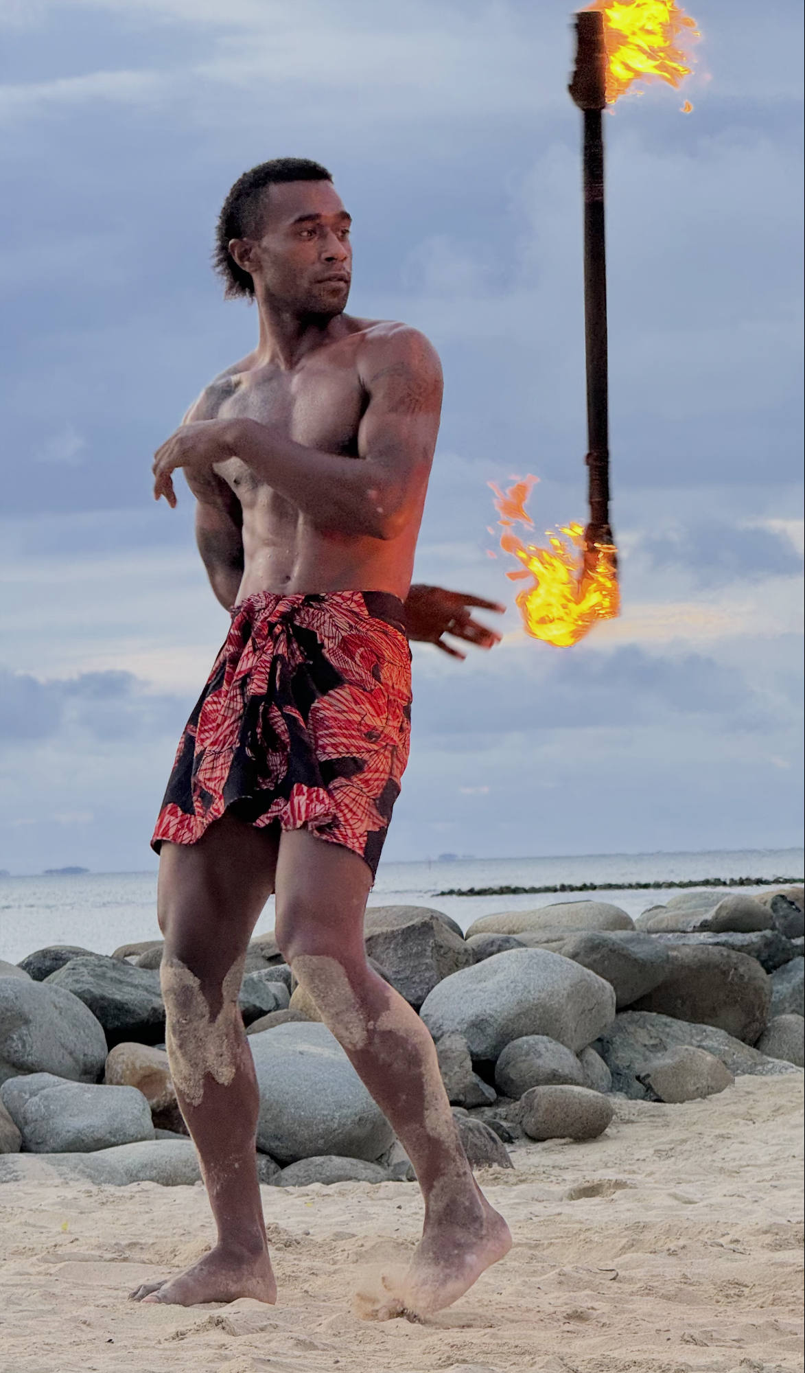 A shirtless man wearing a red and black patterned sarong performs a fire dance on a sandy beach with large rocks in the background. He is captured mid-movement, tossing a double-ended fire staff into the air against a cloudy sky.