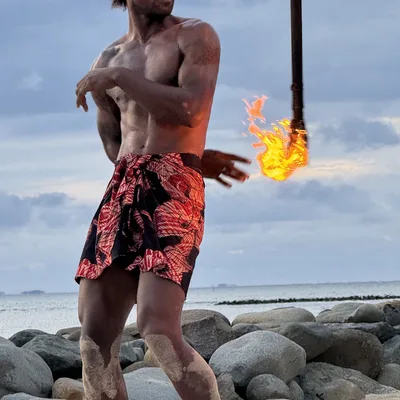 A shirtless man wearing a red and black patterned sarong performs a fire dance on a sandy beach with large rocks in the background. He is captured mid-movement, tossing a double-ended fire staff into the air against a cloudy sky.