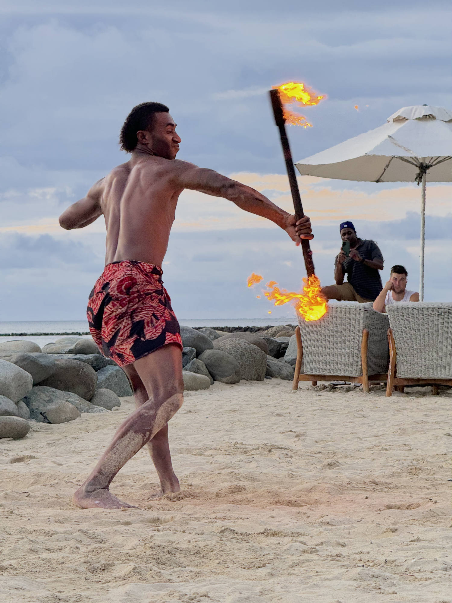 A shirtless male performer in a patterned wrap dances with a double-ended fire staff on a sandy beach. In the background, onlookers watch from patio chairs under a beach umbrella near a rocky shoreline.