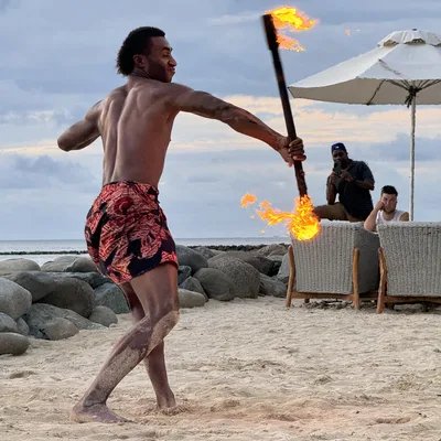 A shirtless male performer in a patterned wrap dances with a double-ended fire staff on a sandy beach. In the background, onlookers watch from patio chairs under a beach umbrella near a rocky shoreline.