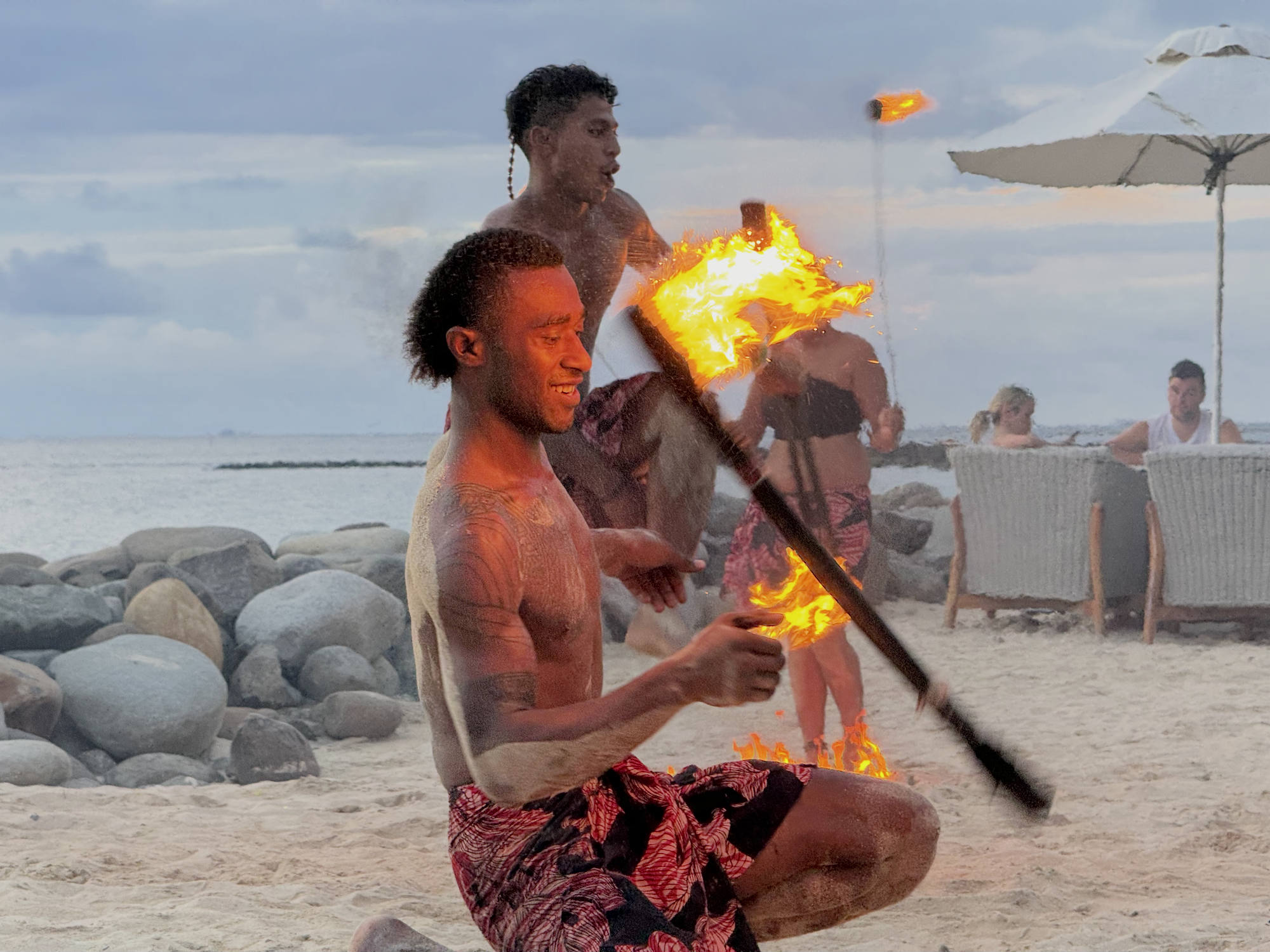 Two shirtless male fire dancers perform with flaming torches on a sandy beach, while spectators watch from chairs under an umbrella in the background.