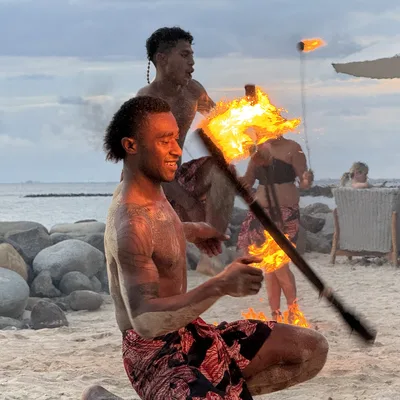 Two shirtless male fire dancers perform with flaming torches on a sandy beach, while spectators watch from chairs under an umbrella in the background.