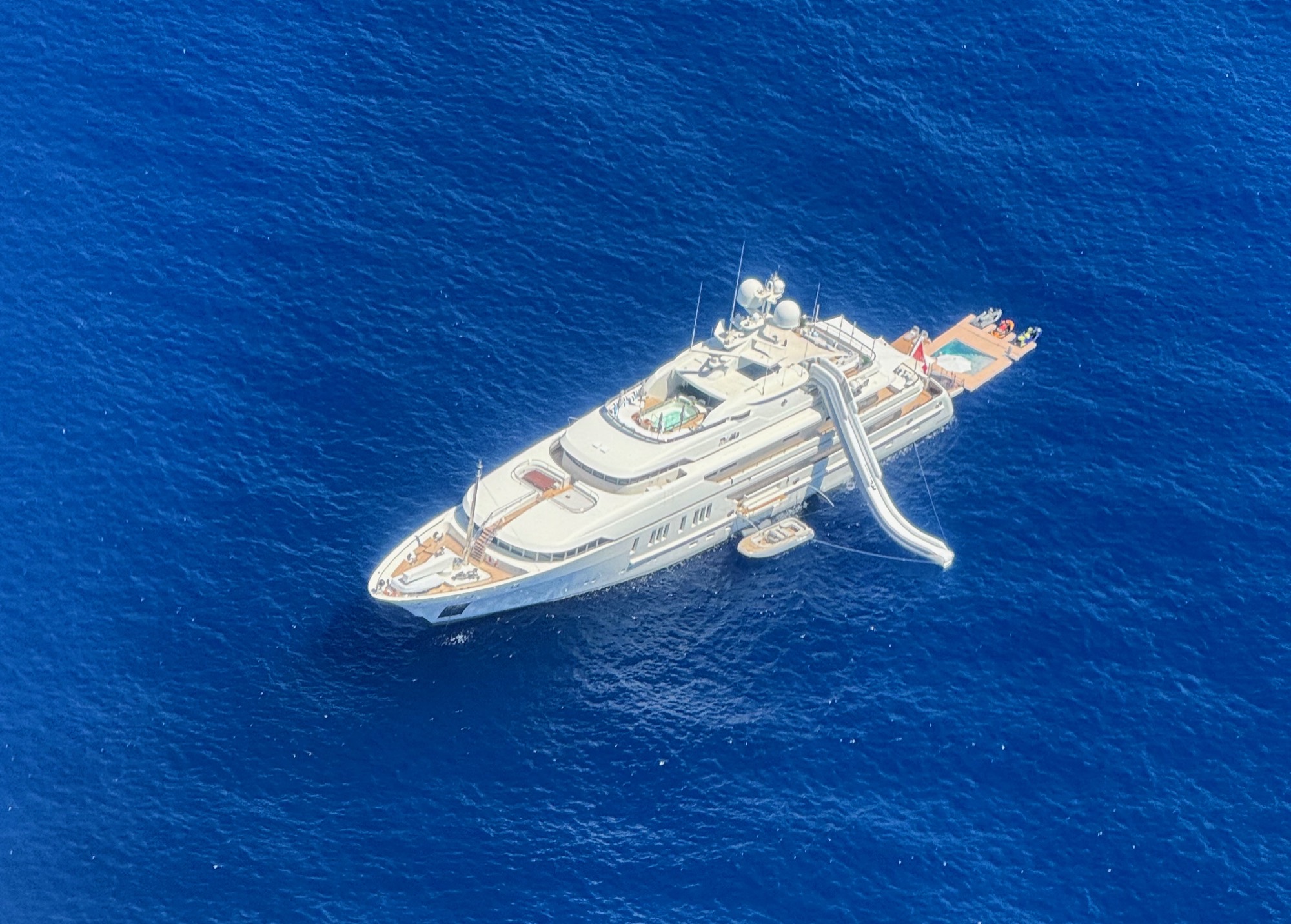 An aerial view shows a large white luxury yacht anchored in deep blue open water. The yacht features an inflatable water slide extending from an upper deck into the sea, a small tender boat tied alongside, and a swim platform at the stern.