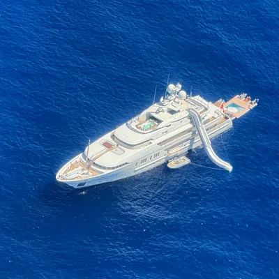 An aerial view shows a large white luxury yacht anchored in deep blue open water. The yacht features an inflatable water slide extending from an upper deck into the sea, a small tender boat tied alongside, and a swim platform at the stern.