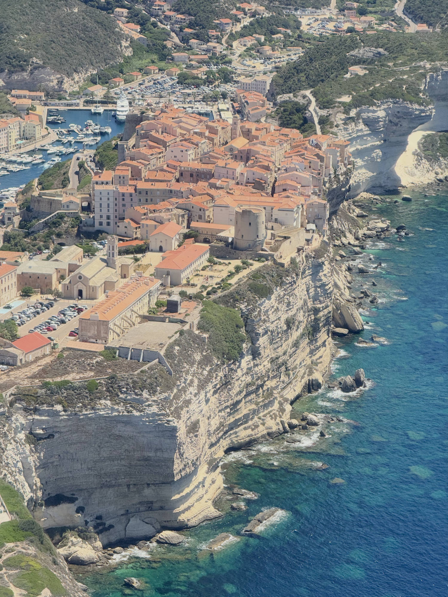 This high-angle aerial photograph captures a coastal town with orange-roofed buildings situated on the edge of steep, white limestone cliffs overlooking a vibrant blue sea. A harbor filled with boats is visible on the left side of the town, while the rugged coastline stretches along the right.