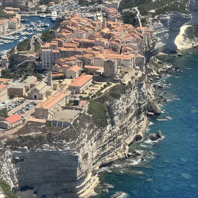 This high-angle aerial photograph captures a coastal town with orange-roofed buildings situated on the edge of steep, white limestone cliffs overlooking a vibrant blue sea. A harbor filled with boats is visible on the left side of the town, while the rugged coastline stretches along the right.