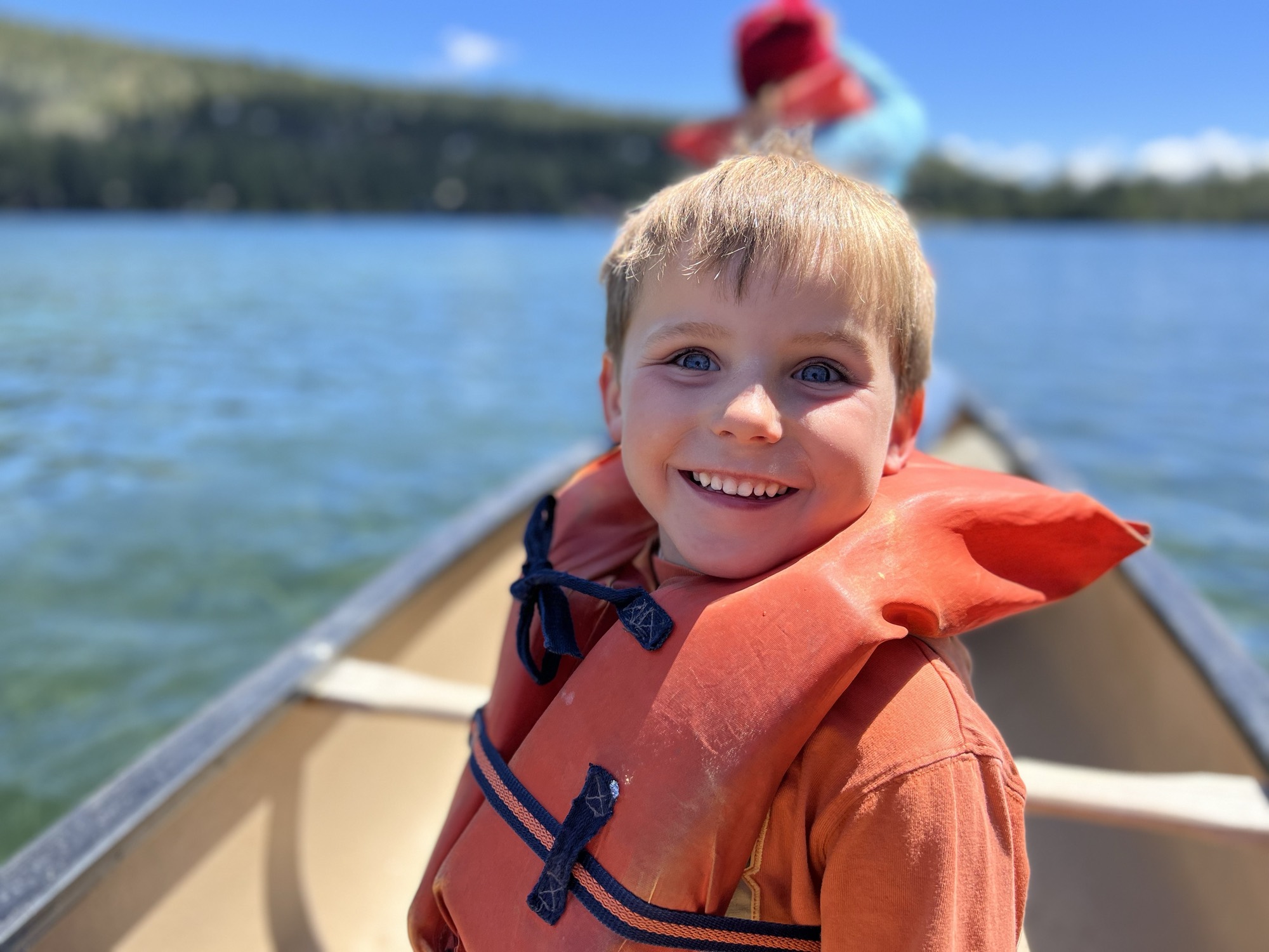 A young boy wearing an orange life jacket smiles at the camera while riding in a canoe on a clear, blue lake. In the blurred background, another person sits in the front of the canoe with forested hills visible under a bright sky.