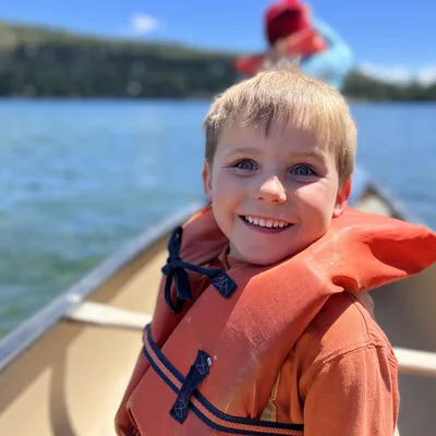 A young boy wearing an orange life jacket smiles at the camera while riding in a canoe on a clear, blue lake. In the blurred background, another person sits in the front of the canoe with forested hills visible under a bright sky.