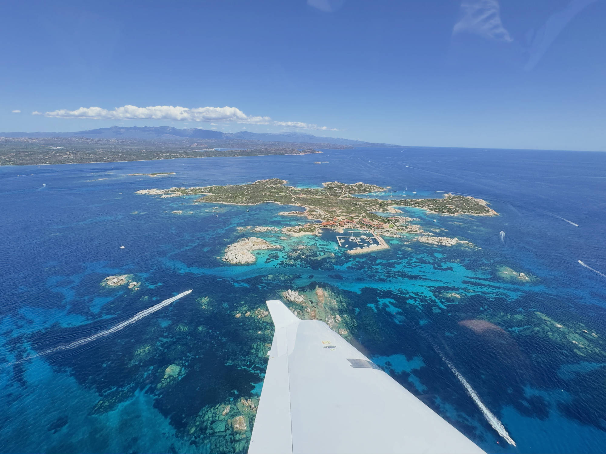 An aerial view from an airplane window shows the aircraft's wing tip pointing toward a small island with a marina, surrounded by vibrant turquoise and deep blue ocean waters. Several boats leave white wakes in the water below, with a larger coastline and mountains visible in the distance under a clear blue sky.