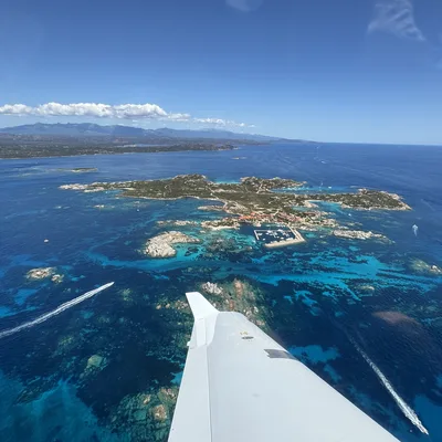 An aerial view from an airplane window shows the aircraft's wing tip pointing toward a small island with a marina, surrounded by vibrant turquoise and deep blue ocean waters. Several boats leave white wakes in the water below, with a larger coastline and mountains visible in the distance under a clear blue sky.