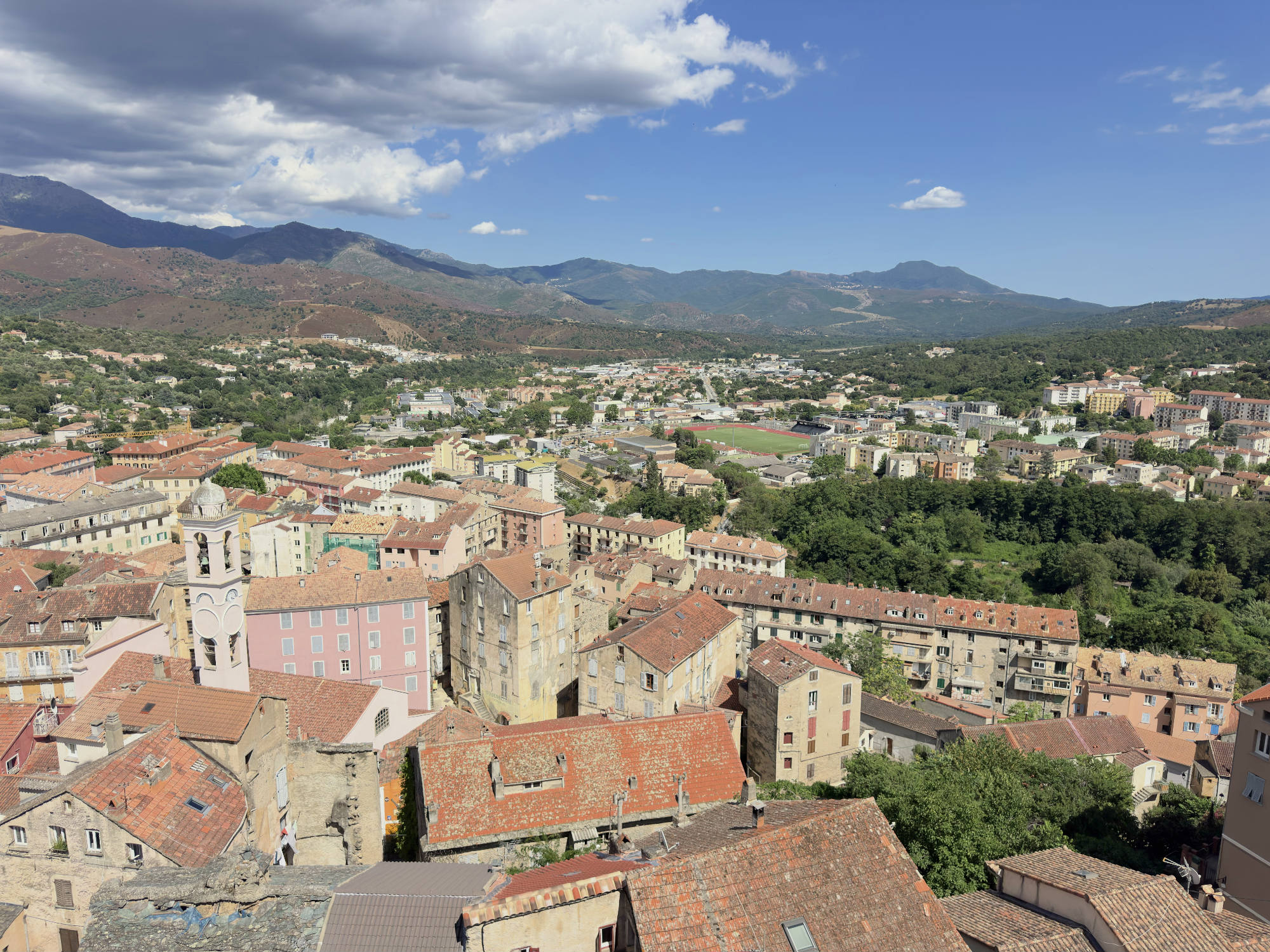 This high-angle, wide shot captures a scenic view of a historic European town with terracotta-tiled roofs, featuring a prominent church tower on the left. In the background, a lush valley with a sports field transitions into rolling green hills and distant mountains under a partly cloudy blue sky.