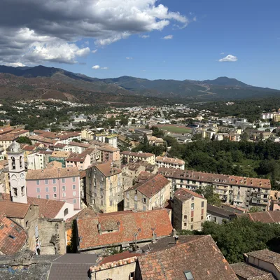 This high-angle, wide shot captures a scenic view of a historic European town with terracotta-tiled roofs, featuring a prominent church tower on the left. In the background, a lush valley with a sports field transitions into rolling green hills and distant mountains under a partly cloudy blue sky.
