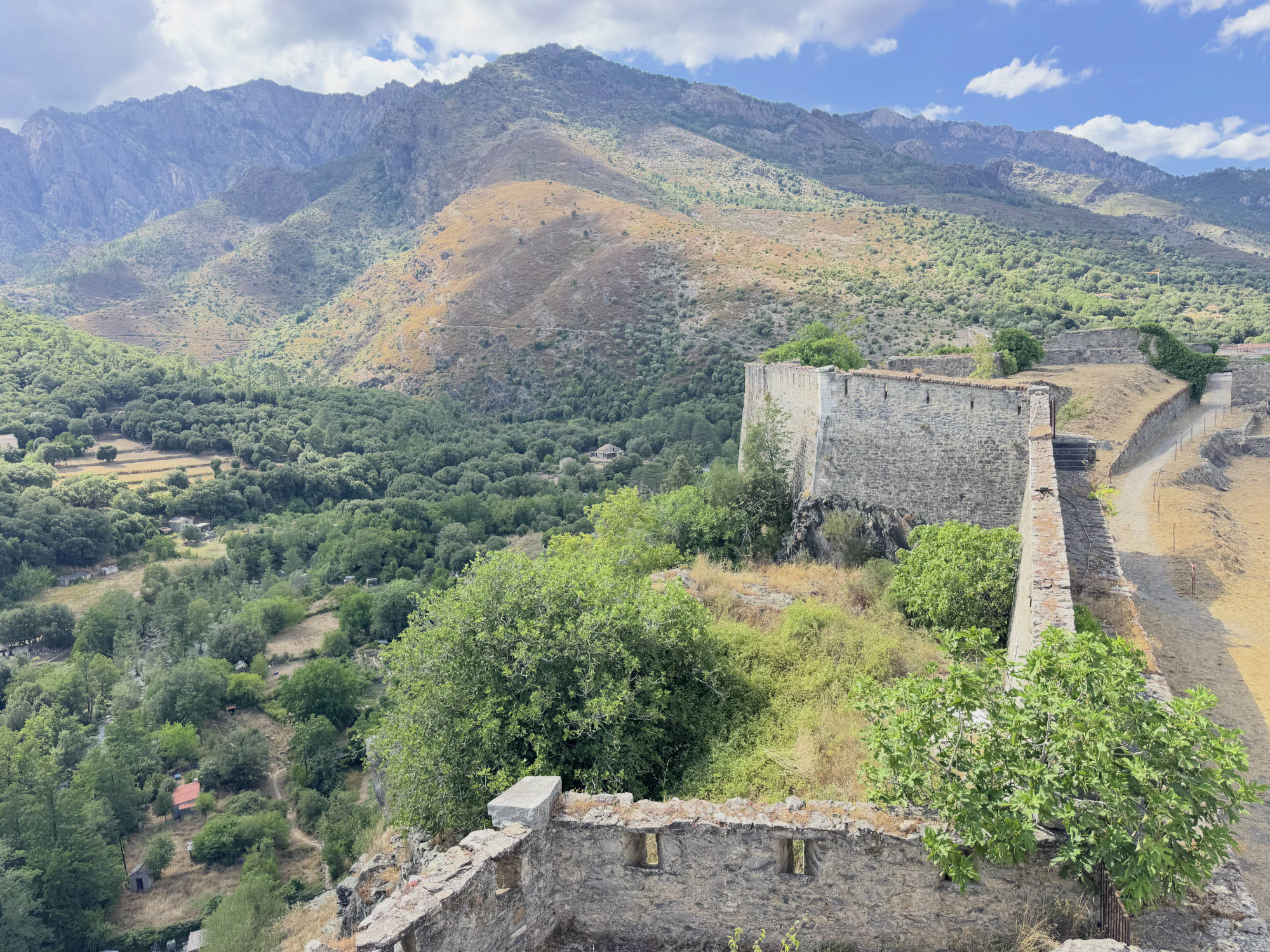 This high-angle shot features the stone walls of a historic fortress overlooking a lush green valley and rugged mountains under a partly cloudy sky. The stone ruins are surrounded by trees and dry grass, with a dirt path visible alongside the main wall.