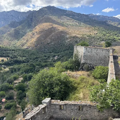 This high-angle shot features the stone walls of a historic fortress overlooking a lush green valley and rugged mountains under a partly cloudy sky. The stone ruins are surrounded by trees and dry grass, with a dirt path visible alongside the main wall.