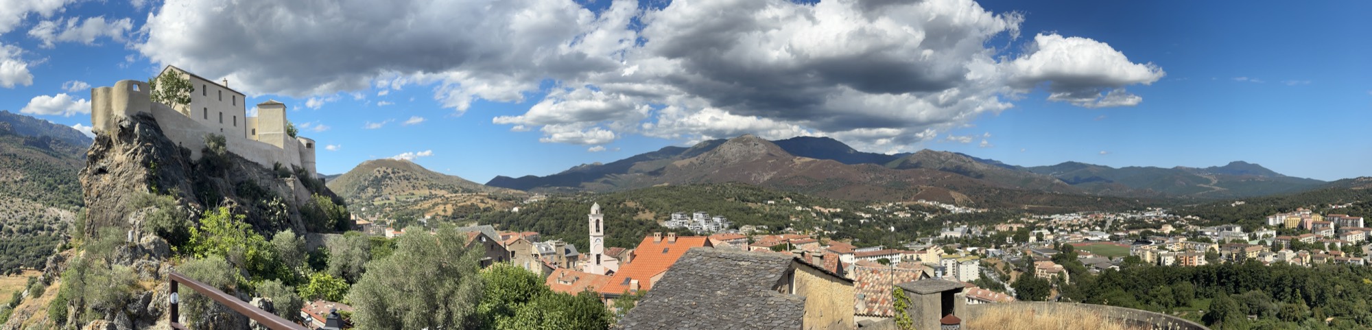 This panoramic photograph captures a scenic view of a hillside town with a large stone citadel perched on a rocky cliff to the left. In the background, rolling mountains stretch across the horizon under a partly cloudy blue sky.