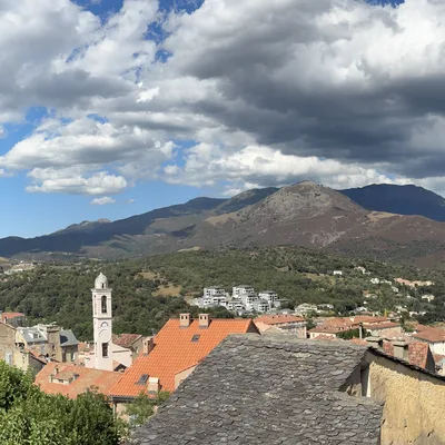 This panoramic photograph captures a scenic view of a hillside town with a large stone citadel perched on a rocky cliff to the left. In the background, rolling mountains stretch across the horizon under a partly cloudy blue sky.