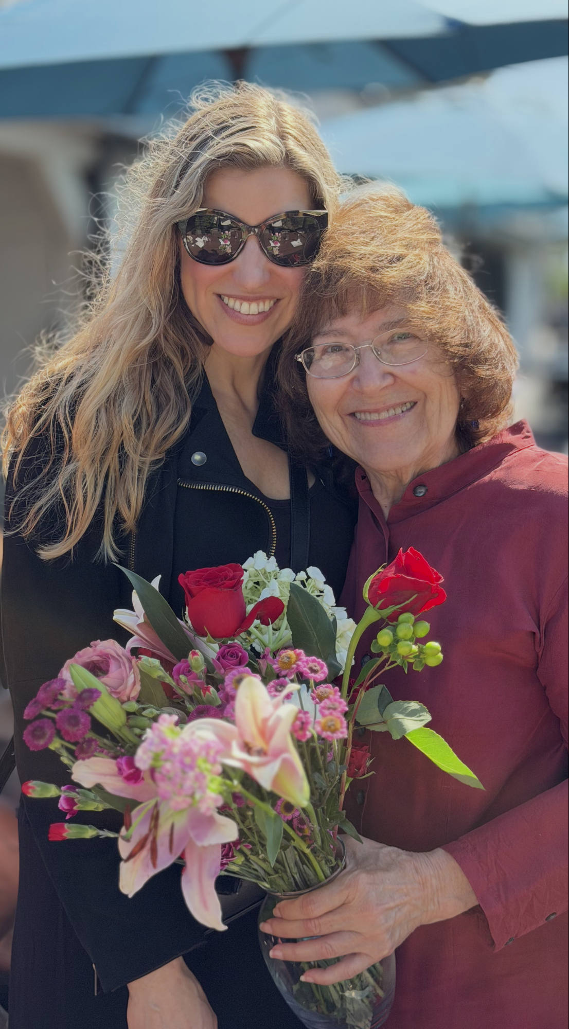 An older woman holding a colorful bouquet of flowers smiles alongside a younger woman wearing sunglasses in an outdoor setting. They are standing close together and smiling for the camera under a blue umbrella.