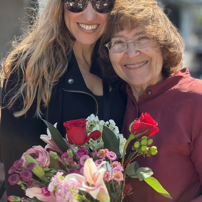 An older woman holding a colorful bouquet of flowers smiles alongside a younger woman wearing sunglasses in an outdoor setting. They are standing close together and smiling for the camera under a blue umbrella.