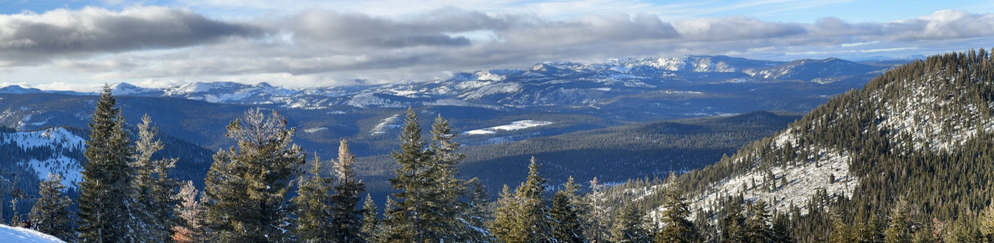 This panoramic photograph captures a vast mountain range covered in snow and dense evergreen forests under a cloudy blue sky. Pine trees dusted with snow frame the foreground, overlooking a deep valley and distant snow-capped peaks.