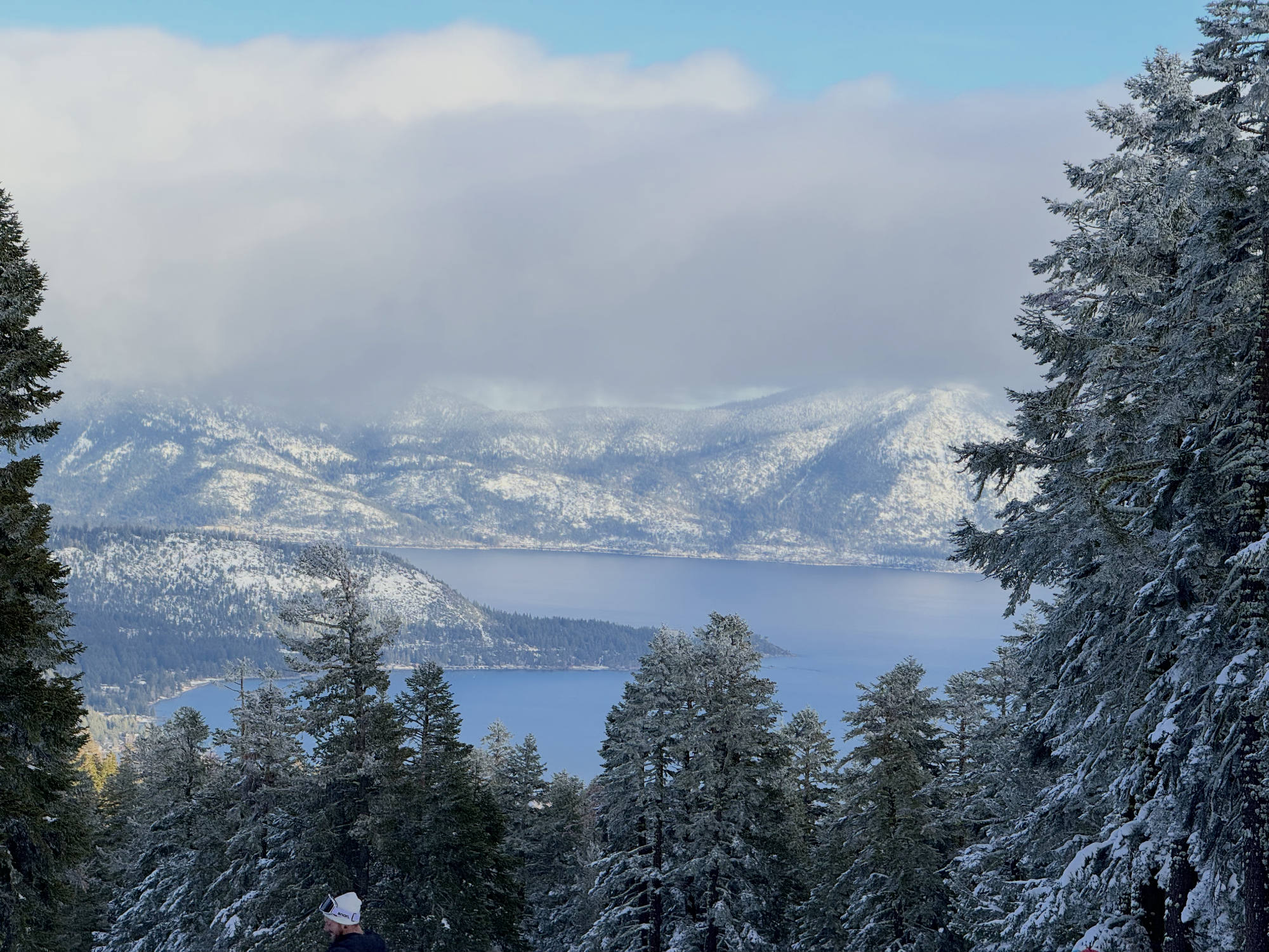 A high-angle view captures a large blue lake surrounded by snow-dusted mountains under a cloudy sky, framed by snow-covered evergreen trees in the foreground. A person wearing a white winter hat is partially visible at the bottom of the frame.