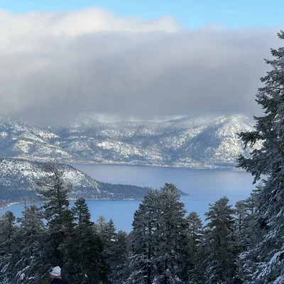 A high-angle view captures a large blue lake surrounded by snow-dusted mountains under a cloudy sky, framed by snow-covered evergreen trees in the foreground. A person wearing a white winter hat is partially visible at the bottom of the frame.
