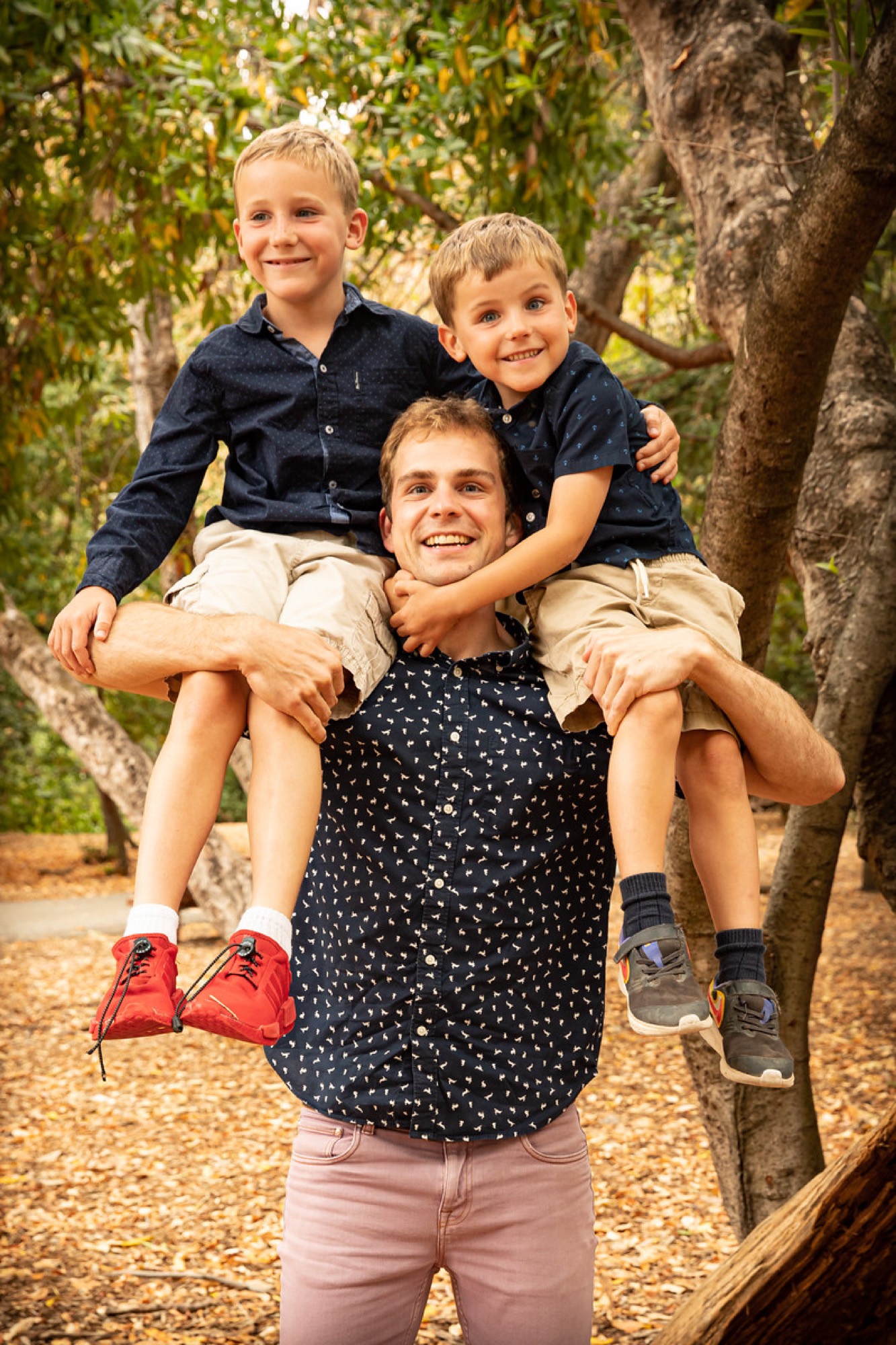 A smiling man stands in a wooded area with two young boys sitting on his shoulders. The group is posing for a portrait amidst trees and fallen leaves.